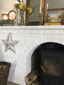 distressed star on white painted fireplace with logs and wood burner and mantle adorned with daffodils candles books and gilt mirror and frames