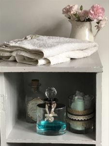 pink flowers in cream jug with white towel and glass bottles and rattan storage inside grey painted pot cupboard