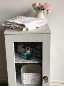 pink flowers in cream jug with white towel and glass bottles and rattan storage inside grey painted pot cupboard with chicken wire front and vintage glass handle