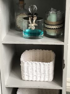 pretty glass bottles and white rattan storage basket on shelves inside grey painted pot cupboard