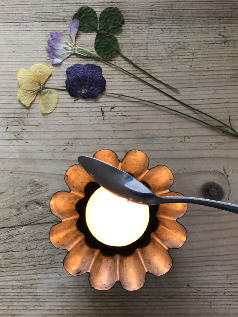 flat lay of tealight with teaspoon over flame and several dried flower heads for pressed flower candle craft