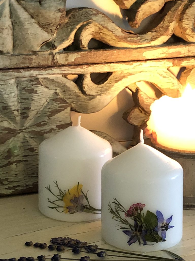 two short white pressed flower candles with ornate wooden backdrop and glowing candle