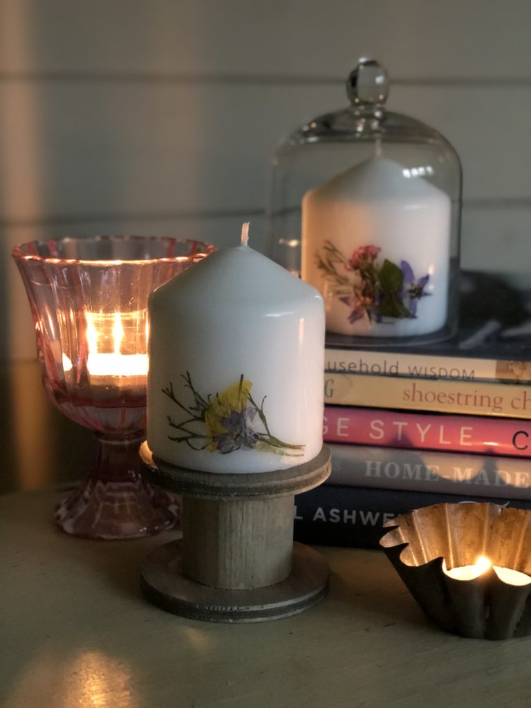 two white pressed flower candles, wooden spool, stack of books, cloche and other candles