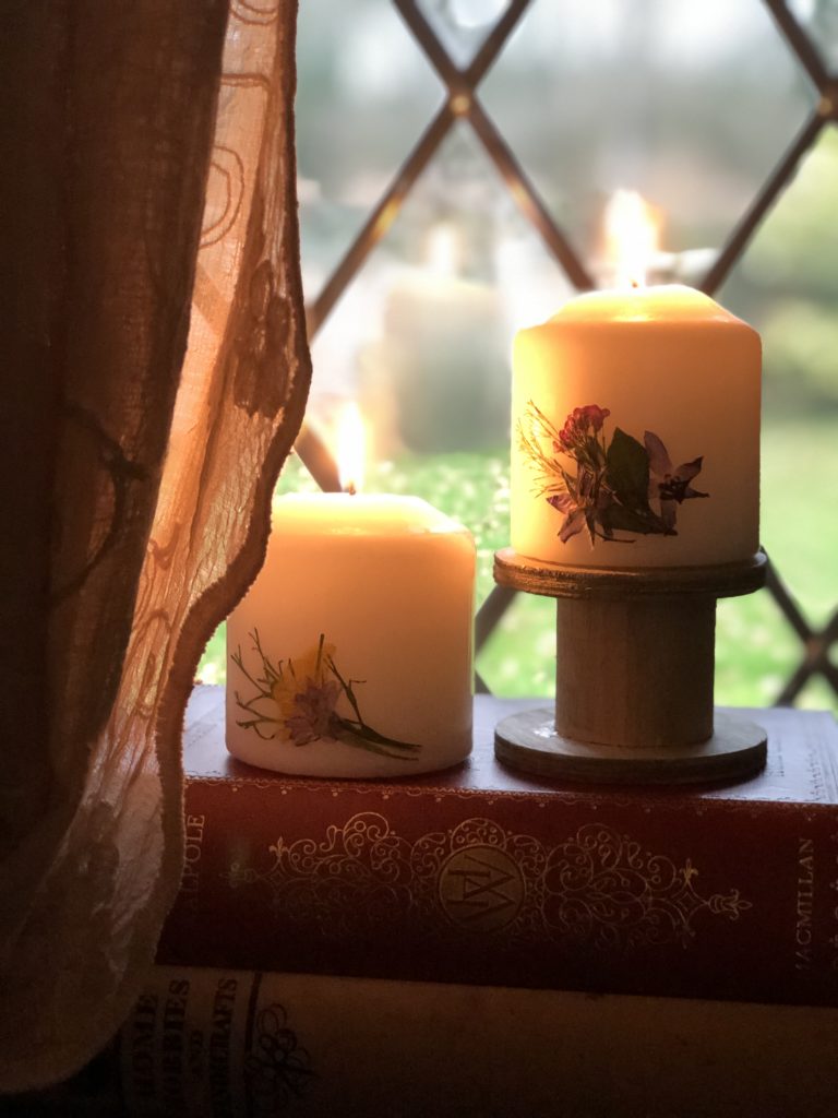 two pressed flower candles glowing in dusk window on book stack and lace curtain detail