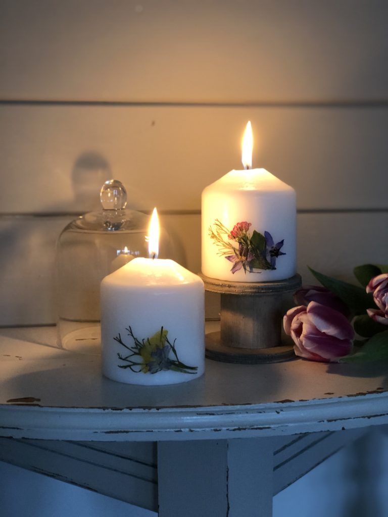 two pressed flower candles glowing on chippy white table with tulips and cloche