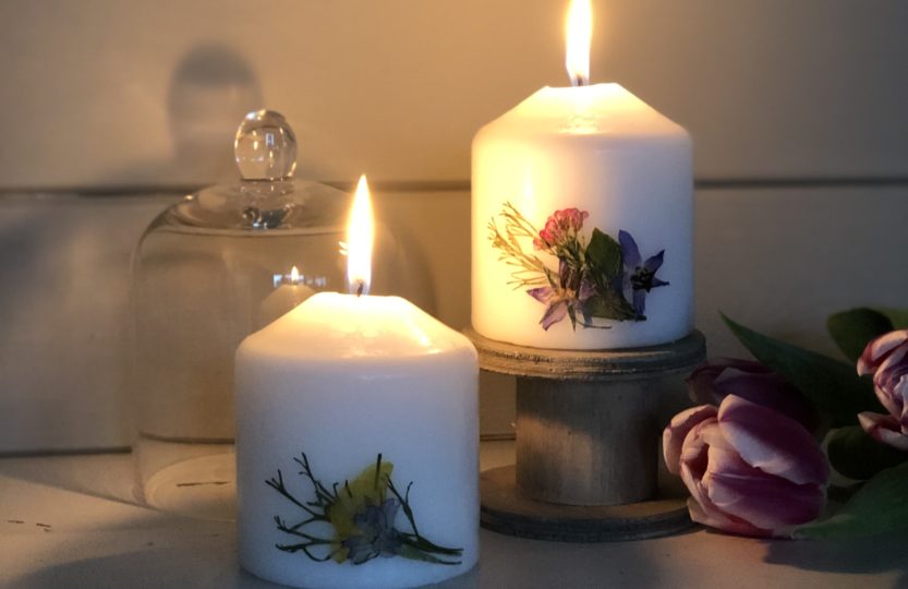 two pressed flower candles glowing on chippy white table with tulips and cloche