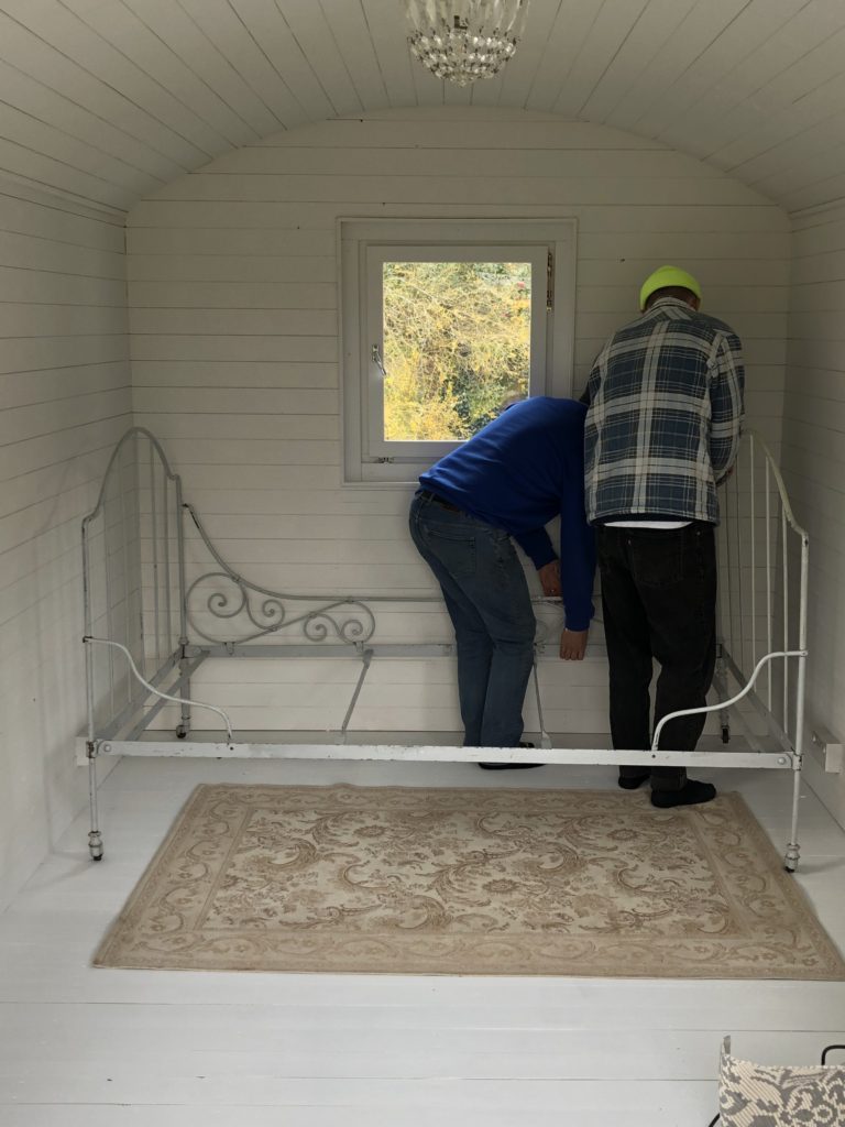 Two men assembling wrought iron bed in white painted shepherd's hut with rug