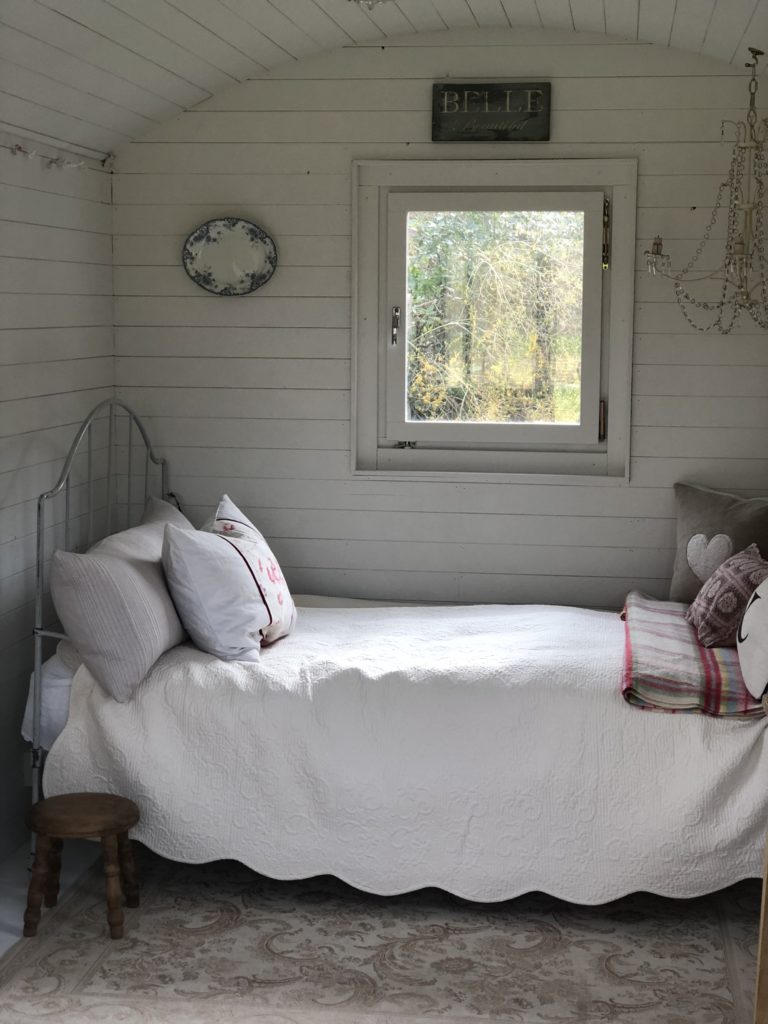 vintage dressed iron bed with cushions and blankets plate on wall and window in white painted shepherd's hut