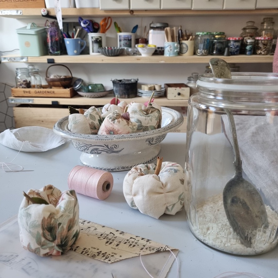 fabric apples, rice, thread, bowls on linen table in craft room with shelves in rear of photo heavily laden with craft accessories and materials