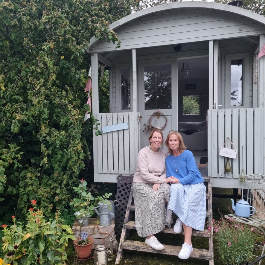 two smiling ladies sit on steps of pretty Shepherds Hut in Cottage Garden