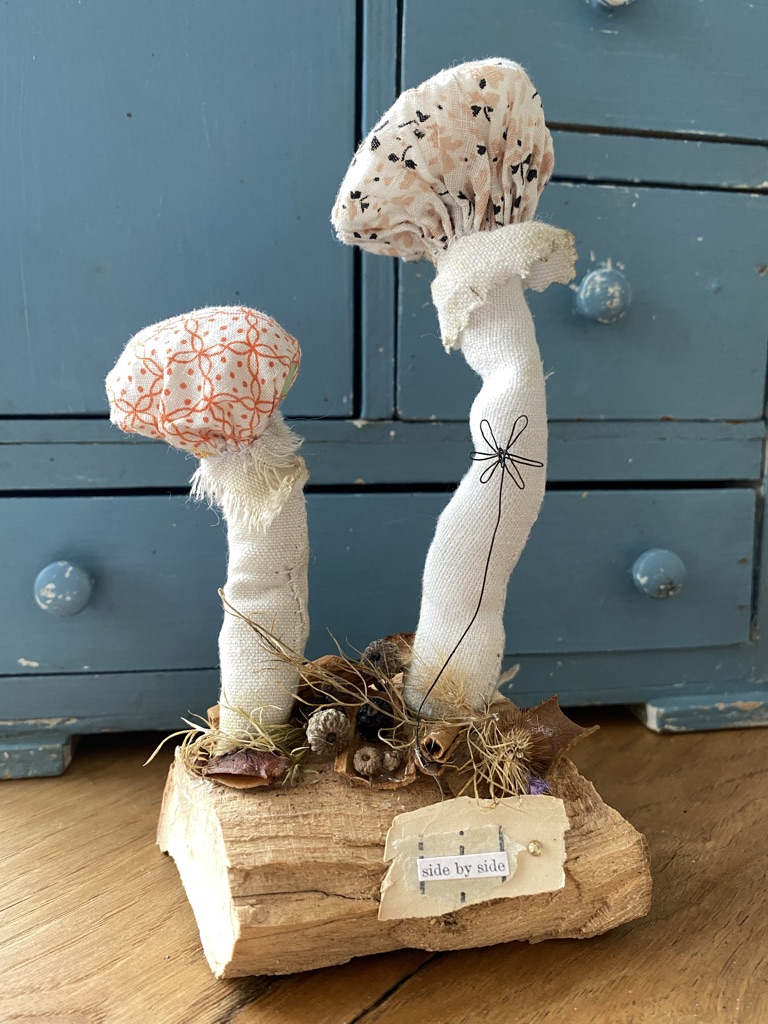 a craft woodland setting of two textile fabric mushrooms on top of wood with berries and dried flowers at the base, a wire flower and a paper quote in front of a teal chippy cupboard