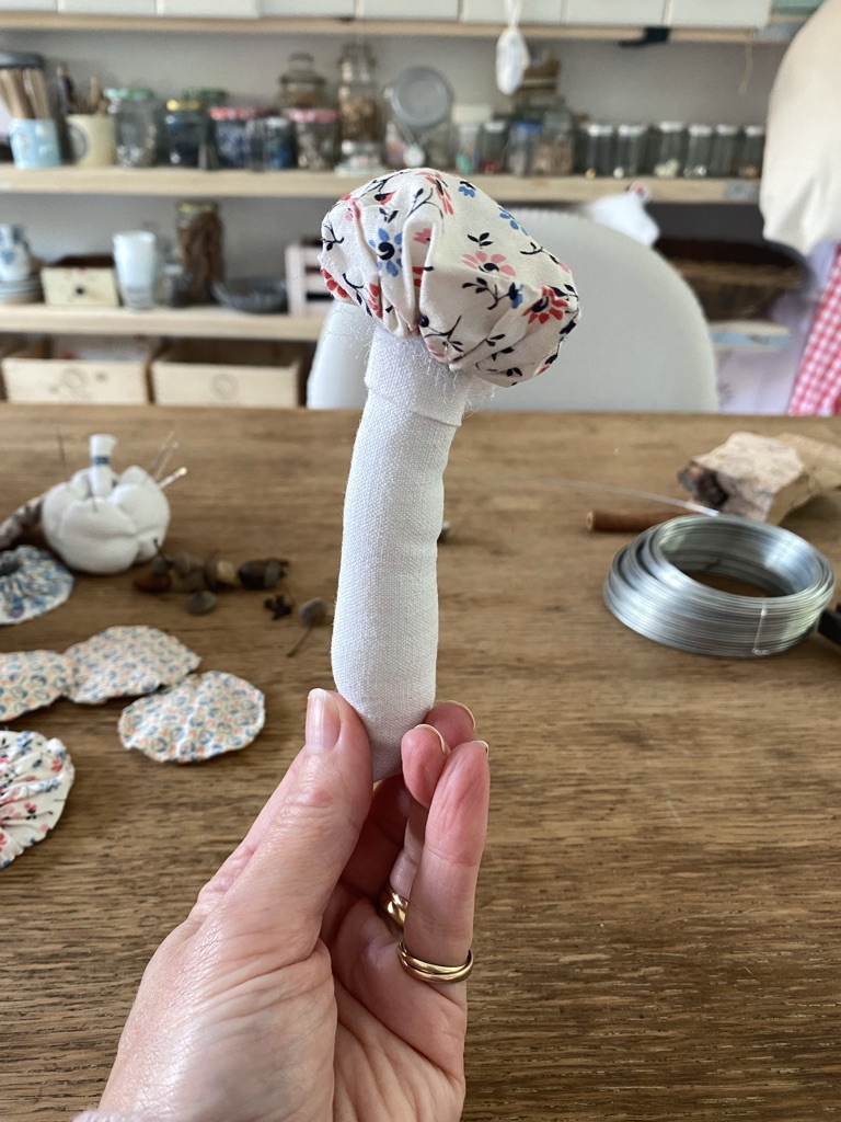 hand holds a textile fabric mushroom in a craft room with suffolk puffs and wire on wooden craft table