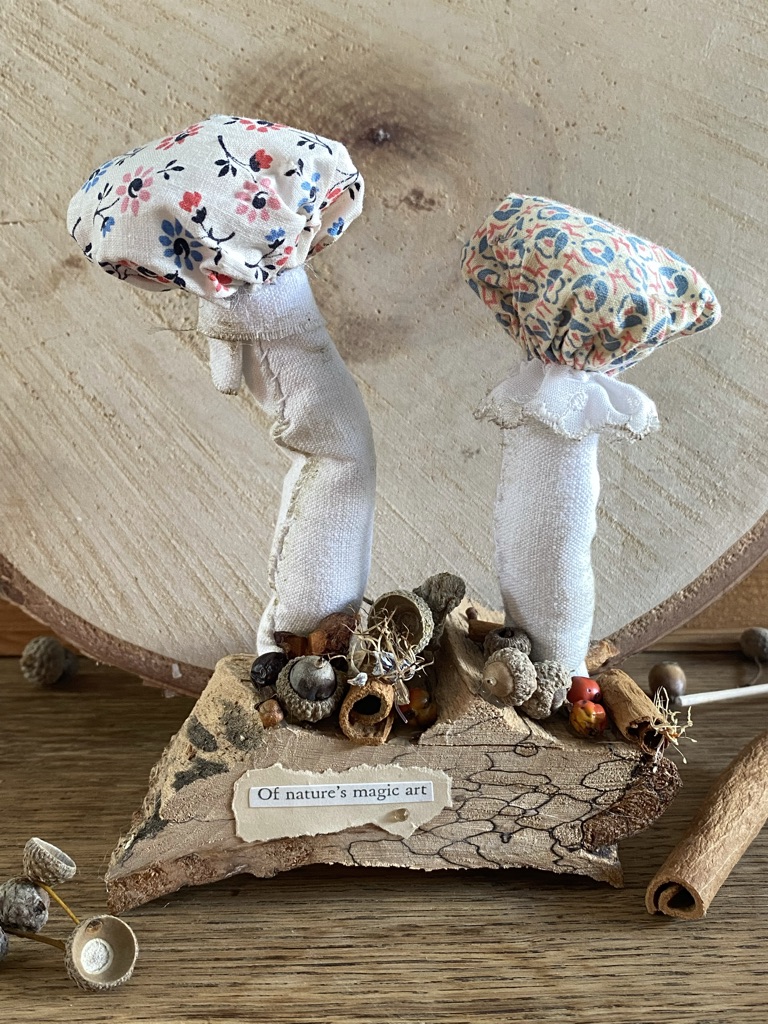 a craft woodland setting of two textile fabric mushrooms on top of wood with berries and dried flowers at the base and a paper quote