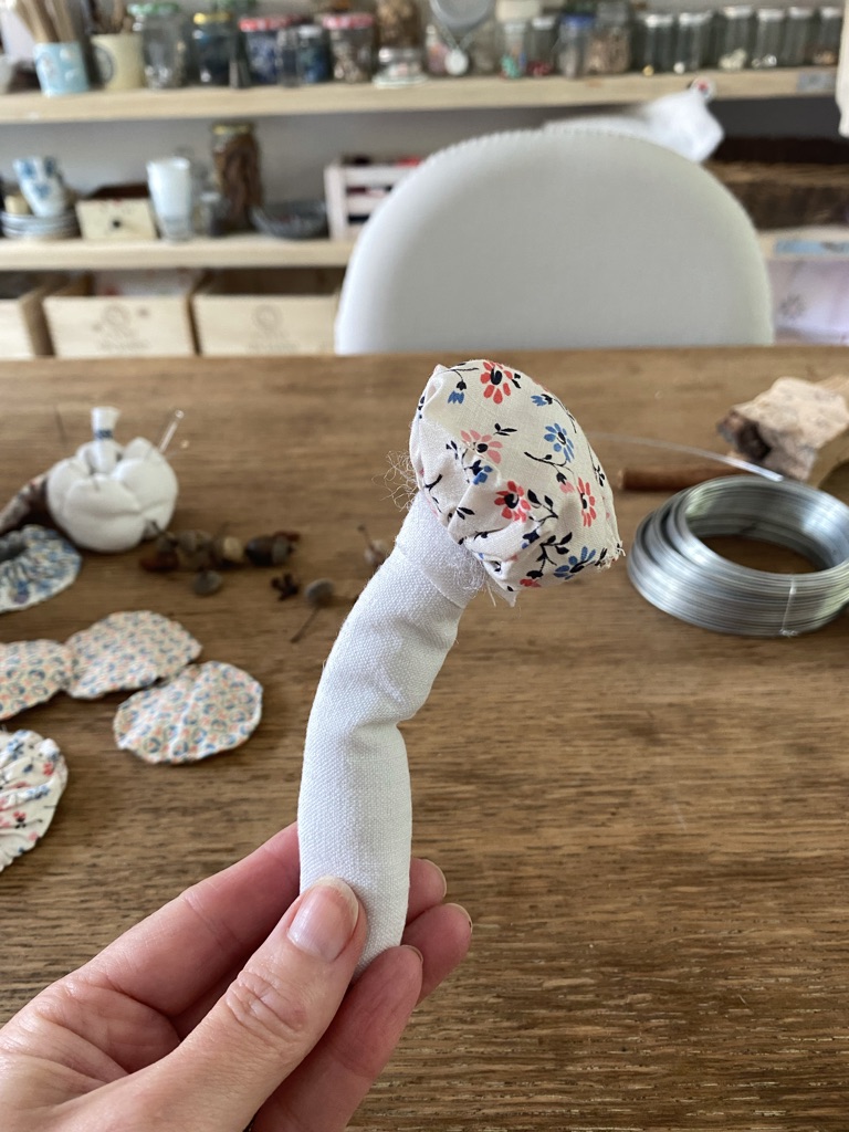 hand holds a textile fabric mushroom in a craft room with suffolk puffs and wire on wooden craft table