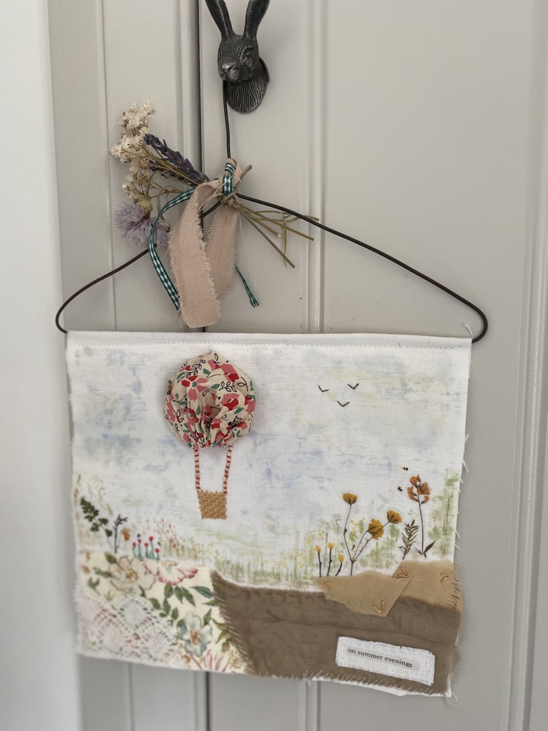 a mixed media fabric collage wall hanging decoration of suffolk puff yo-yo hot air balloon in countryside setting complete with rusty wire hanger