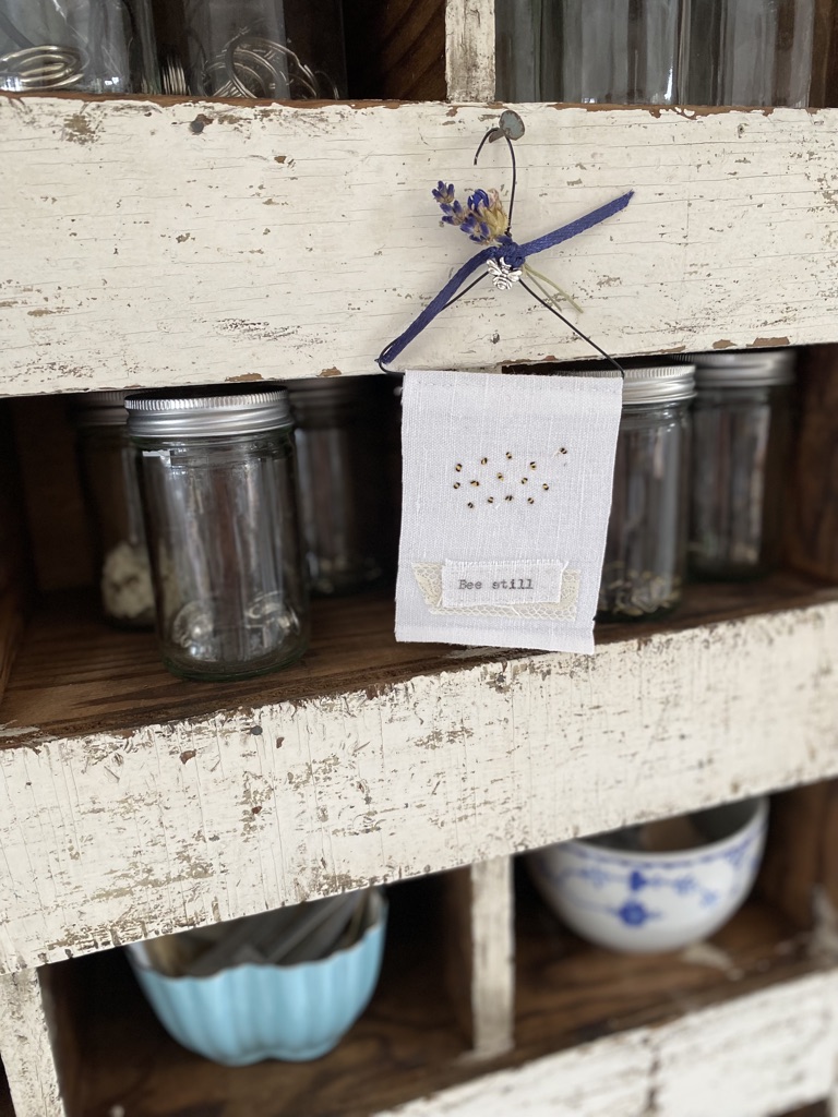 A simple bumble bee embroidery hangs from off white vintage chippy cubby holes full of vintage pots and glass jars with trinkets 