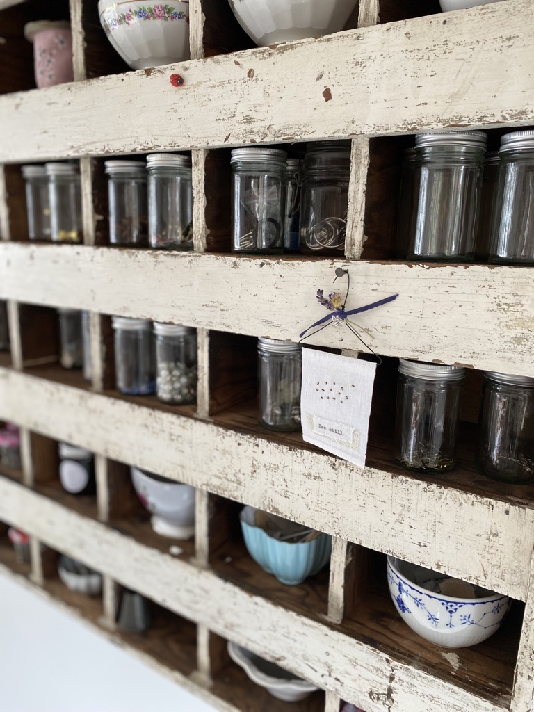 A simple bumble bee embroidery hangs from off white vintage chippy cubby holes full of vintage pots and glass jars with trinkets 